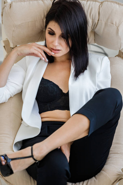 Studio portrait of beautiful brunette woman wearing black pants, white jacket and black bra lying in armchair at home interior  - Фото, изображение