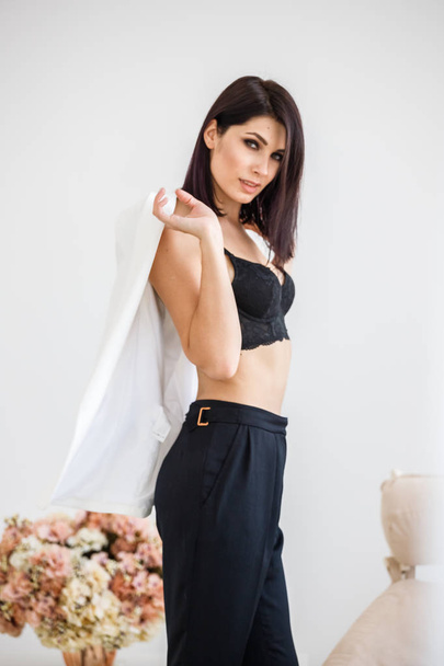 Studio portrait of beautiful brunette woman wearing black pants, white jacket and black bra standing at home interior with flowers bouquet on background  - Foto, Imagem
