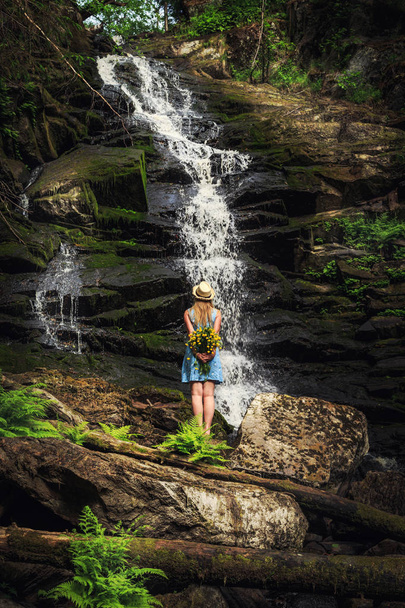 Woman in hat with bouquet of flowers near a forest waterfall. Feeling of relaxation and freedom. - Foto, afbeelding
