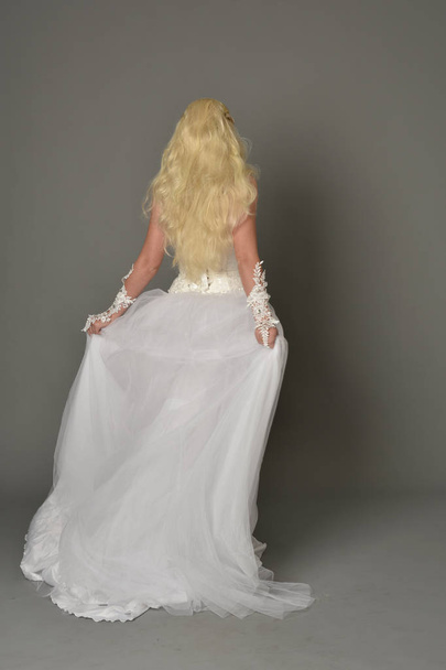 full length portrait of blonde girl wearing white gown, standing pose with back to the camera. grey studio background. - Фото, зображення