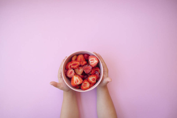 Kinderhände mit einer rosafarbenen Schale voller schöner frischer saftiger Erdbeeren auf pastellrosa Hintergrund mit Textfläche. - Foto, Bild