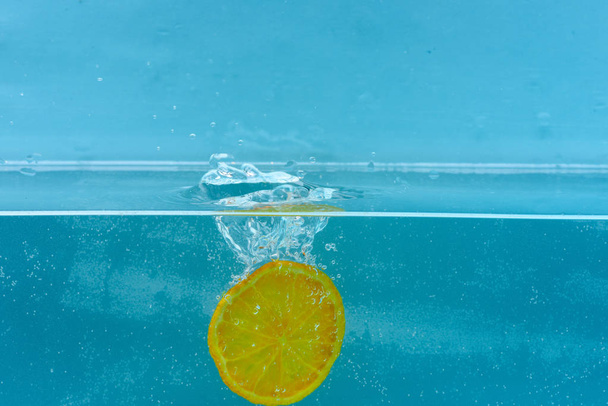 Slice of orange under water with transparent bubbles and water drops splashes. Fruit fall into water, blue background. Cocktail making concept. Orange in pure, transparent liquid - Fotografie, Obrázek