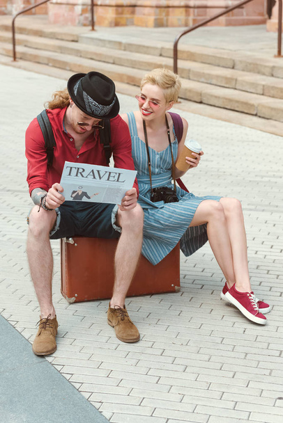 couple of tourists with coffee to go reading travel newspaper and sitting on suitcase  - Fotó, kép
