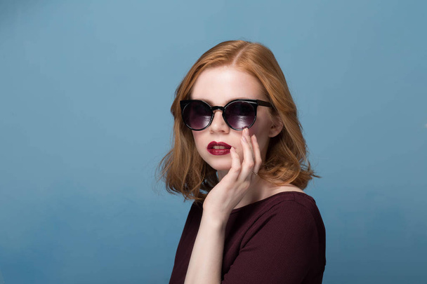 beautiful redhead girl in a maroon blouse red lipstick bright makeup in black sunglasses on a blue background closeup portrait - Photo, Image