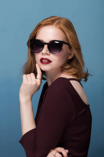 beautiful girl with makeup and red lips in the Burgundy jacket in sunglasses on blue background profile of the hand on the chin, open mouth - Фото, зображення