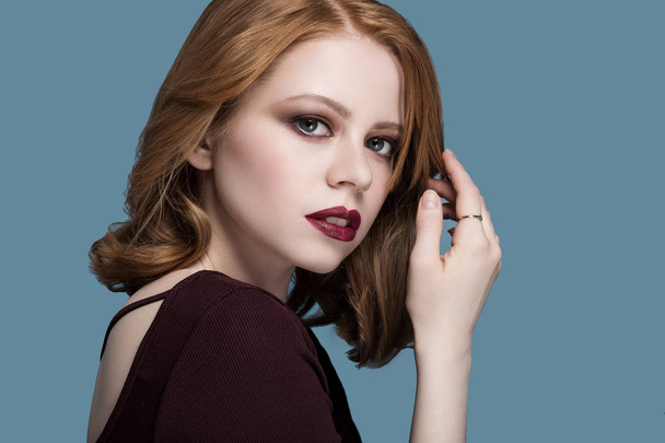 beautiful sexy redhead girl with fresh, bright makeup grey big eyes in a Burgundy sweater over blue background hands in hair looking into the camera lips parted red lips contour - Photo, Image