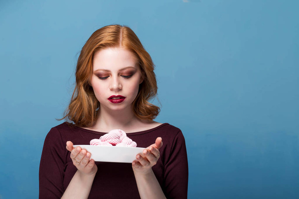 Pretty young woman posing with zephyr. Looking at marshmallows in a bowl. Blue background, copy space - Foto, Imagen