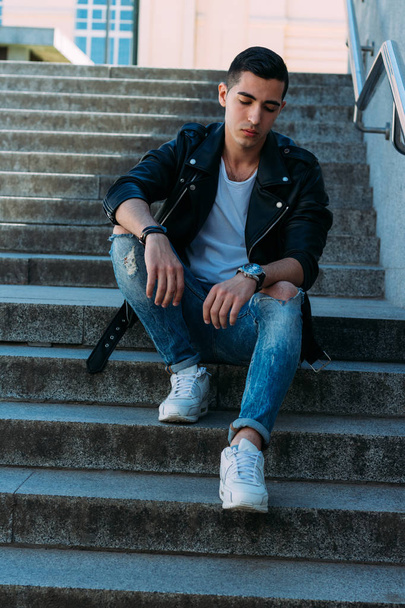 Sexy man posing sits on the steps near railing. an attractive young man In a black leather jacket, a white T-shirt and white sneakers. watch on hand - Фото, зображення