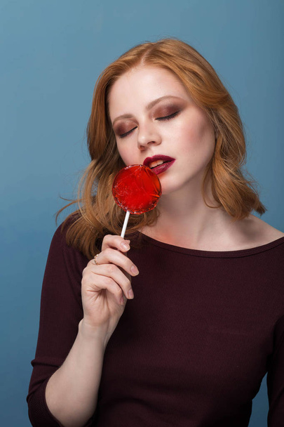 Stylish female model posing with lollipop and with closed eyes on blue background. - Foto, afbeelding