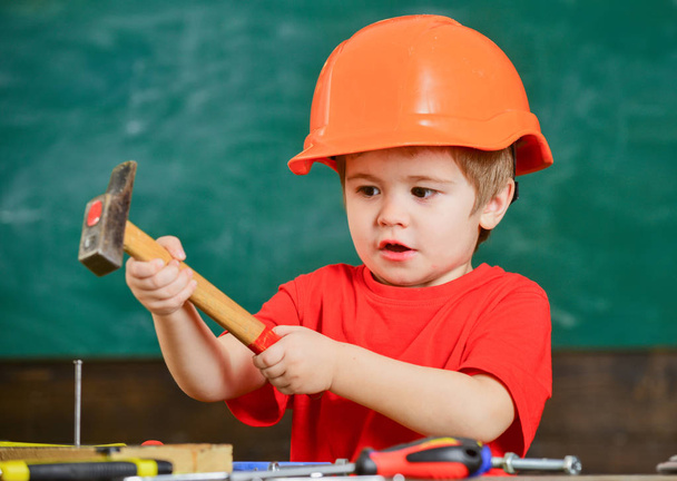 Excited kid playing with heavy hammer. Cute boy in orange helmet helping in workshop. Future profession concept - Foto, afbeelding