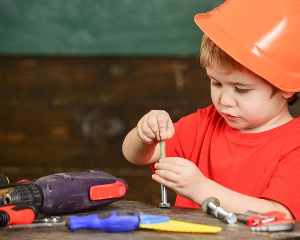 Toddler on busy face plays with bolts at home in workshop. Handcrafting concept. Child in hard hat, helmet playing with hex bolts as builder or repairer, handcrafting. Kid boy play as handyman - 写真・画像