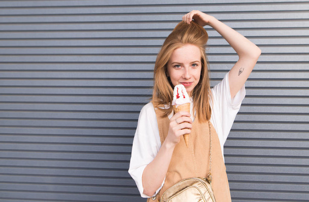 Mignon élégant ore girl pose sur un fond gris avec de la crème glacée dans ses mains. Portrait de fille rousse avec des taches de rousseur et dessert à la main sur le mur de fond
. - Photo, image