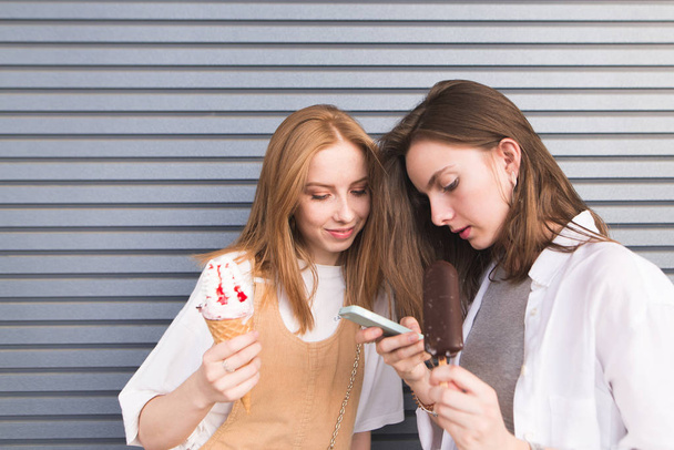 Çekici genç kızlar dondurma ellerinde bir smartphone ile gri bir duvar arka plan üzerinde duruyor. Arka plan duvar üzerinde şirin kız portresi. - Fotoğraf, Görsel
