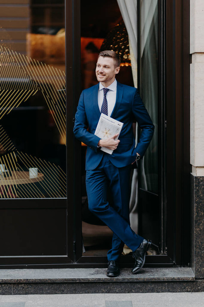 Confident successful businessman in formal suit, keeps hand in pocket, holds business magazine, stands near entrance of cozy cafeteria. People, job and occupation concept - Fotó, kép