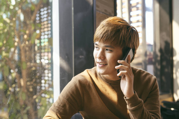 close-up retrato de sorrindo jovem asiático homem falando no celular e sentado à mesa vazia com café borrado interior no fundo
 - Foto, Imagem