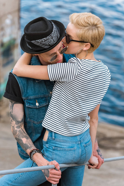 boyfriend with tattoos and stylish girlfriend hugging and leaning on railing near river - Фото, изображение