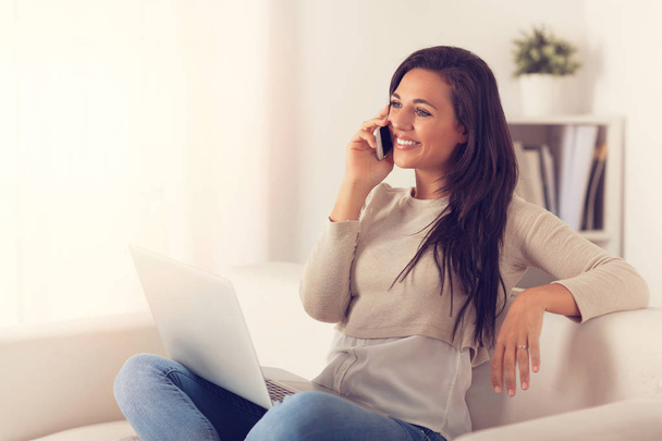 Portrait of smiling woman using laptop while speaking on the phone sitting on a couch  - Photo, Image