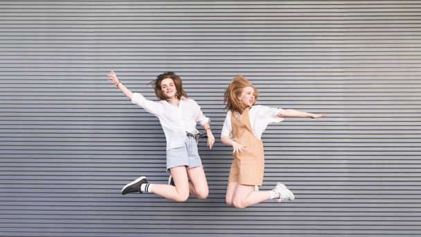 Two stylish happy girls jump together on a gray background. Portrait of smiling fashionable girlfriends in motion that jump on a dark background. - Foto, afbeelding