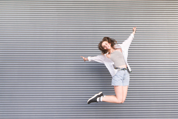 Retrato de uma menina bonita feliz que salta em roupas leves no fundo de uma parede cinza. Jovens atraentes para saltar sobre um fundo cinza
. - Foto, Imagem
