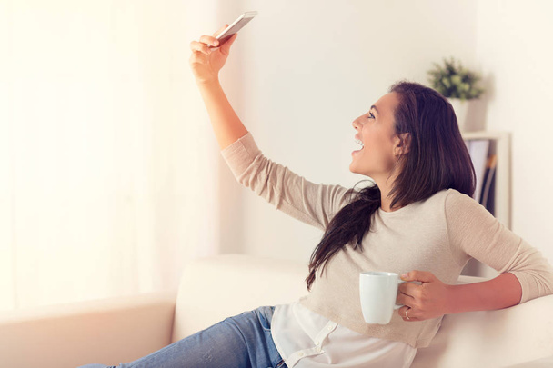 Portrait de jeune femme prenant un autoportrait avec le téléphone intelligent sur un canapé à la maison
  - Photo, image