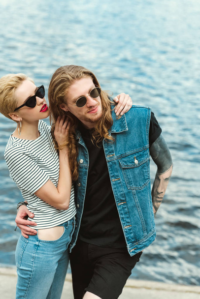 boyfriend with tattoos and stylish girlfriend cuddling on bridge near river - Zdjęcie, obraz