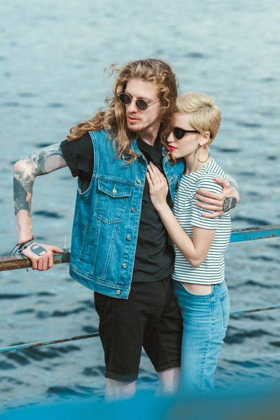 boyfriend with tattoos and stylish girlfriend hugging on bridge near river - Фото, зображення