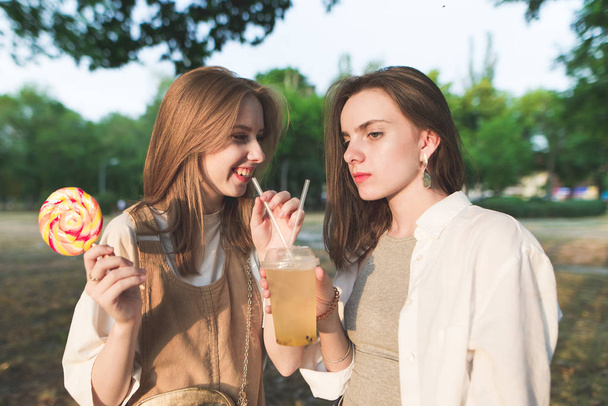 Portret van twee mooie meisjes in het park met lollies en limonade in de hand. Twee stijlvolle jonge vrienden zijn permanent in het park en chatten. Ontspan met een vriendin buitenshuis op een zomeravond. - Foto, afbeelding
