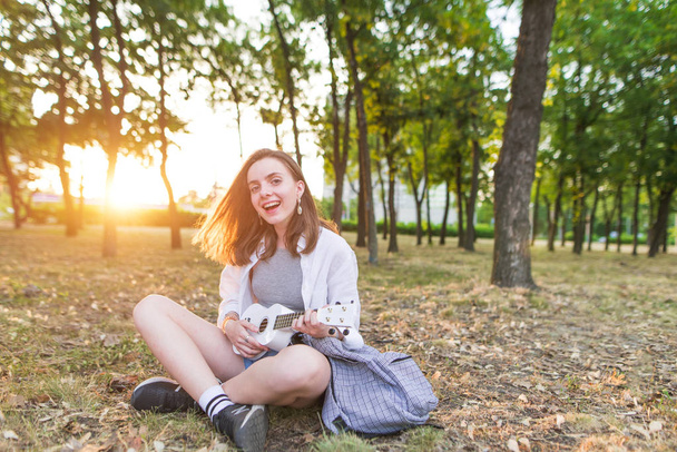 sonriente chica con estilo se sienta en un parque, juega en un ukelele blanco y mira a la cámara. La chica feliz es un músico tocando ukelele y canta al aire libre
. - Foto, Imagen