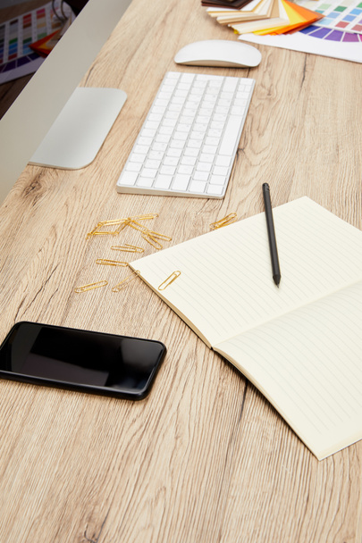 Close-up van ontwerper werkruimte met smartphone, lege notebook, beeldscherm en toetsenbord op houten oppervlak - Foto, afbeelding