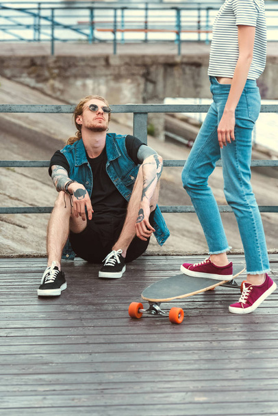 cropped image of boyfriend with tattoos sitting on bridge and girlfriend standing on skateboard - Fotó, kép