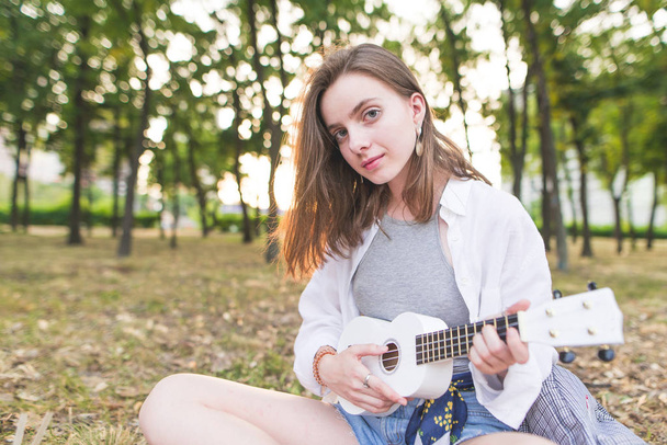 Portrait of an attractive girl sitting in a park with ukulele in her hands and looking into the camera. Pretty young girl plays the ukulele in the park. - Photo, image
