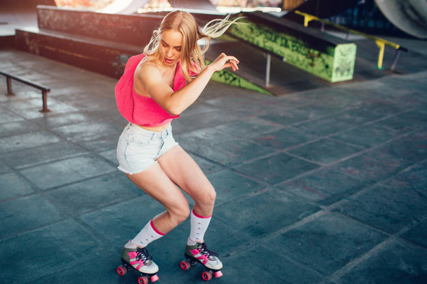 Beautiful blonde girl is doing some tricks during rollerblading. She is standing in a squat position and looking down. Her hands are aside of body. She is exercising in training room. - Photo, image