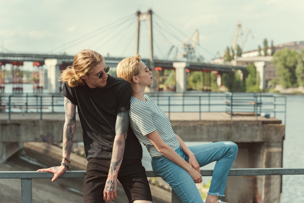 boyfriend with tattoos and stylish girlfriend touching with backs and sitting on railing of bridge - 写真・画像