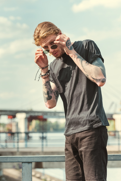 όμορφος μοντέρνος άνδρας τατουάζ φορώντας γυαλιά ηλίου στη γέφυρα - Φωτογραφία, εικόνα