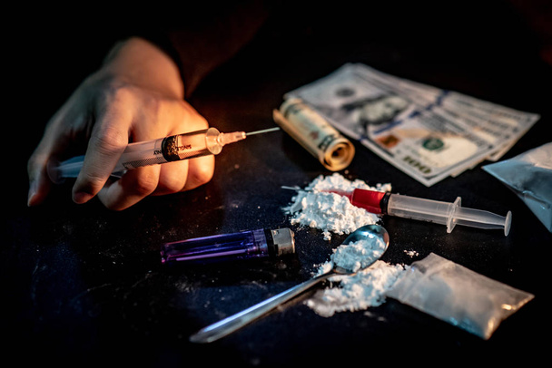 Мужчина-наркоман держит шприц для инъекций наркотиков рядом с героиновым порошком, ложкой и зажигалкой для приготовления героина и деньгами на тёмном полу. Концепция тяжелой наркомании
 - Фото, изображение