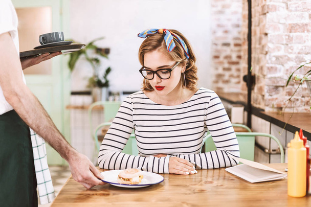 Tarjoilija musta lippis antaa järjestystä mukava nainen silmälasit joka istuu pöydässä ja harkitsee hampurilainen kahvilassa
 - Valokuva, kuva