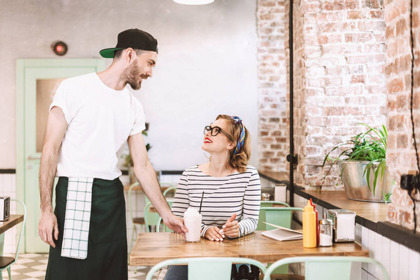 Waiter in black cap giving milkshake to pretty smiling lady in eyeglasses which happily looking at him while sitting at the table in cafe - Foto, Imagen