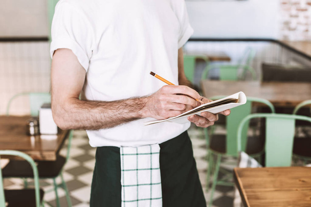 Feche a foto do garçom de pé com lápis e escreva a ordem no caderno enquanto trabalhava no café
 - Foto, Imagem