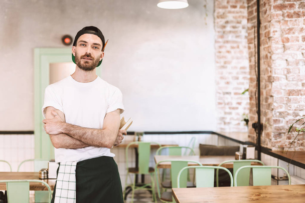Young man in black cap,white t-shirt and apron standing and dreamily looking in camera while working in cafe - Photo, Image