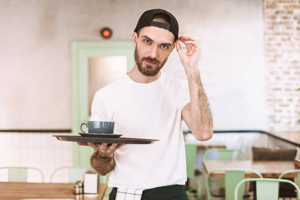 junger Mann mit schwarzer Mütze und weißem T-Shirt steht und hält Tablett mit Tasse Kaffee, während er im Café verträumt in die Kamera schaut - Foto, Bild