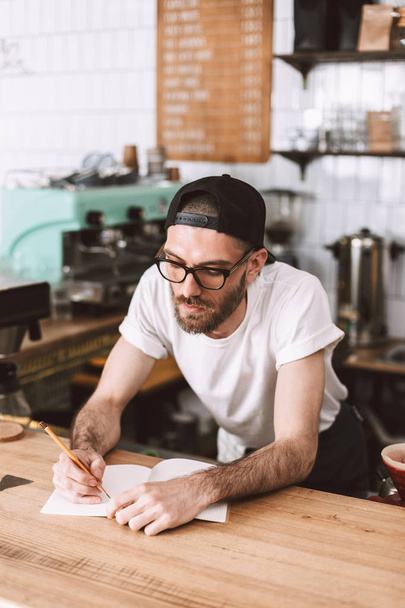 Young pensive man in eyeglasses and cap standing behind bar counter and writing in notepad while working in cafe - Foto, Bild