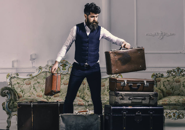 Luggage and travelling concept. Macho elegant on thoughtful face standing near pile of vintage suitcase. Man, traveller with beard and mustache packing luggage before trip, luxury interior background. - Foto, Bild