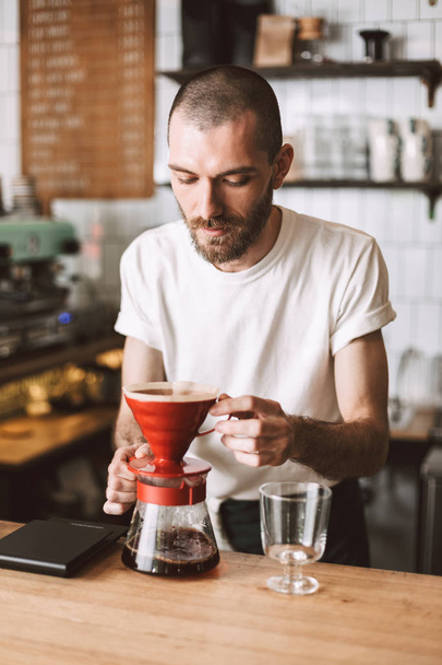 Молодой задумчивый бариста, стоящий у барной стойки и готовящийся наливать кофе во время работы в кафе
 - Фото, изображение