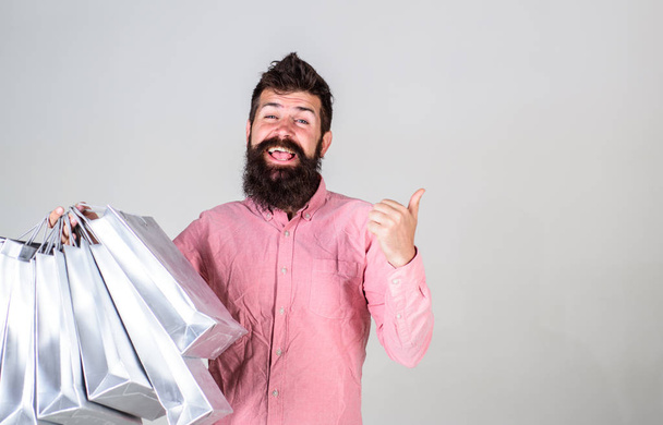 Hipster on smiling face recommends to buy. Man with beard and mustache carries bunch of shopping bags, grey background. Guy shopping on sales season, pointing at blank surface. Recommendation concept - Fotoğraf, Görsel