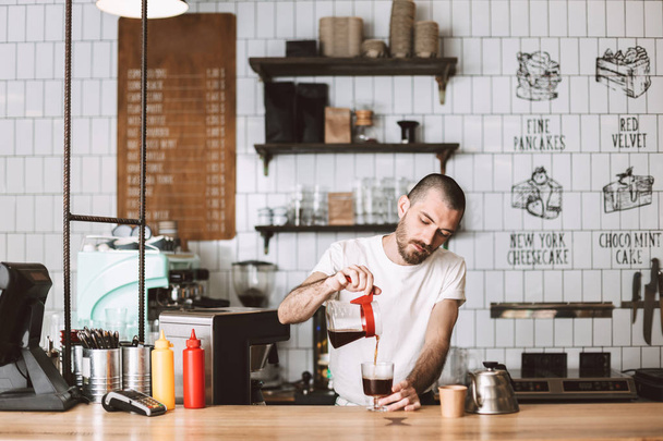 junger Barista steht an der Theke und gießt verträumt Filterkaffee ins Glas, während er im Café arbeitet - Foto, Bild