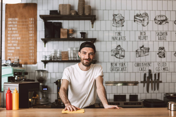 Smiling barista in cap standing and wiping bar counter while happily looking in camera in cafe - Foto, Bild