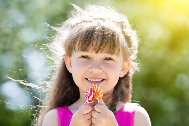 Beautiful little girl is playing with big sweet candy in the city park. A girl in a red dress with sweets in her hands. - Foto, Imagen