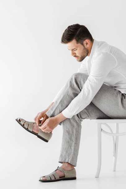 side view of man in linen clothes sitting on chair and putting on sandals isolated on grey background  - Fotó, kép