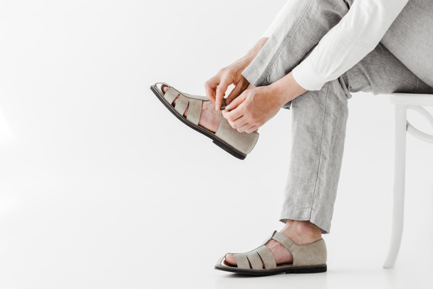 cropped image of male model in linen trousers sitting on chair and putting on sandals isolated on grey background  - Φωτογραφία, εικόνα