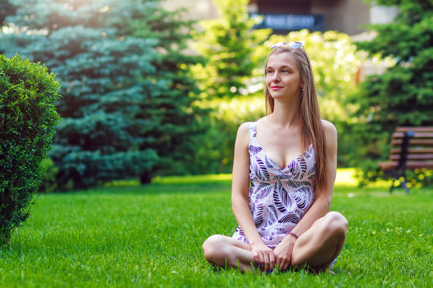 Porträt einer hübschen, jungen, fröhlichen Frau, die auf grünem Gras sitzt und sich ausruht, die Beine gekreuzt - Foto, Bild
